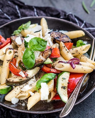 Schwarzer Teller mit Penne, gegrillter Aubergine, Zucchini, Paprika und Basilikumblättern auf dunklem Grund