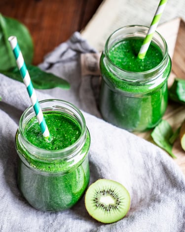 Zwei Gläser mit grünem Smoothie und grün-weißen Strohhalmen neben Kiwihälften auf hellgrauem Tuch