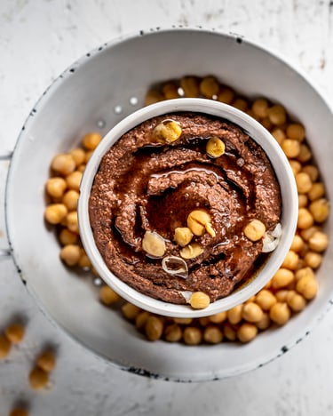 Schokoladen-Hummus mit ganzen Kichererbsen in einer Schale, von oben fotografiert auf hellem Untergrund.
