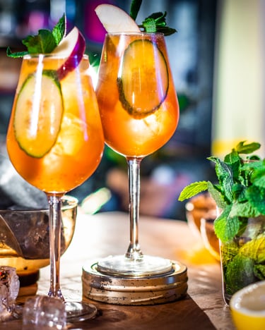 Pimm's Cup in zwei Aperitif-Gläsern gegen das Licht fotografiert. Dekoriert wird der Drink von Gurkenscheiben, die im Licht schimmern. Das Foto wurde vor einer Bar-Szenerie aufgenommen.