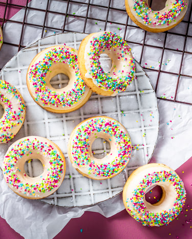 Die Donuts mit bunten Streuseln liegen auf einem pinken Tisch und einem weißen Backpapier verteilt. Daneben steht eine Schüssel mit Eierlikör-Glasur und die bunten Streusel sind drum herum verteilt