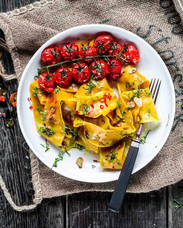 Breite Pasta aglio e olio mit geschmorten Tomaten auf einem weißen Teller, fotografiert auf einem alten Jutesack und auf dunklem Holzuntergrund.