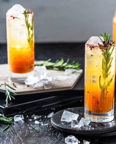 Mit einem schönen Farbverlauf von Orange zu Gelb erstrahlen die Mocktails in den Farben des Sonnenuntergangs. Dekoriert mit einem Stiel Rosmarin und rosa Pfefferkörnern machen sie richtig was her.