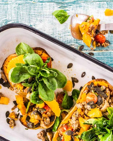Zwei Hälften Butternutkürbis mit Hackfleisch und Feldsalat in weißer Auflaufform auf blauem Holzuntergrund