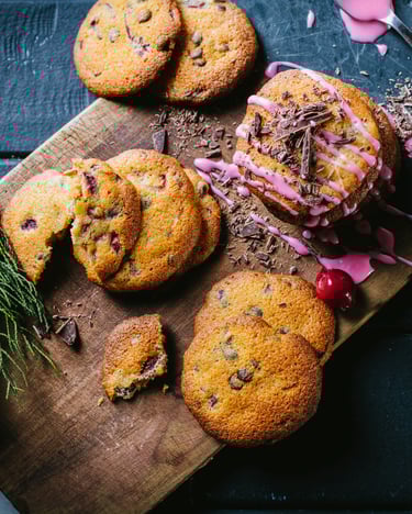 Schwarzwälder Kirschplätzchen mit rosa Zuckerguss auf einem Holzbrettchen mit Tannenzweig, nebenstehend Kirschen in einer Tasse.