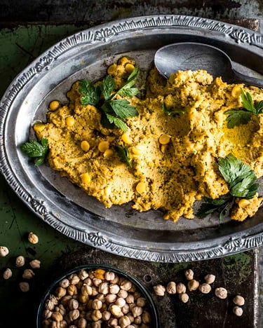Einfacher Hummus, angerichtet auf silbernem, verzierten Teller, mit Löffel auf dem ovalen Teller, fotografiert auf dunklem, rustikalen Untergrund von oben. Kichererbsen sind zu sehen.