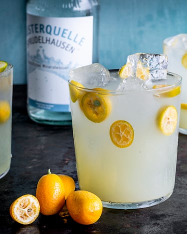 Drei Gläser gefüllt mit Eiswürfeln, Mocktails und halbierten Kumquats auf schwarzem Tisch.