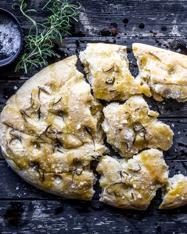 Klassische Focaccia mit Meersalz und Rosmarin auf dunklem Grund. Focaccia ist in kleine, mundgerechte Teile gerupft.