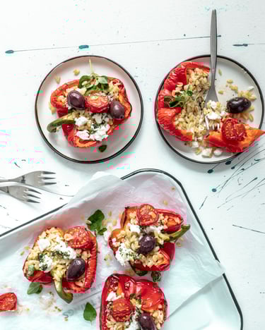 Rote Paprikahälften gefüllt mit Ebli, Tomaten, Oliven und Feta auf weißem Tablett und zwei weißen Tellern angerichtet