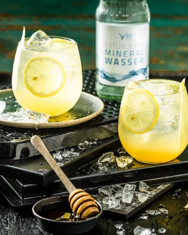 Blass-edel sieht der gesunde Drink aus Ingwer, Zitrone und Essig aus, mit einem leichten Farbverlauf von Gelb zu Transparent. Ein Honiglöffel und ein Ingwerstück liegen bei den zwei Gläsern auf einem schwarzen Untergrund.