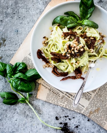 Pasta mit Avocadocreme, bestreut mit Pinienkernen, daneben etwas Basilikum. Von oben fotografiert auf einem Tekller, der auf einer Zeitung steht auf hellem Steinuntergrund.