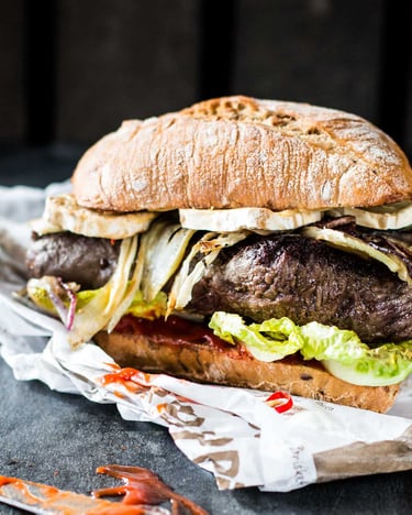 Steakburger mit Brie, Fenchel und Chili auf Zeitungspapier und Steinuntergrund.
