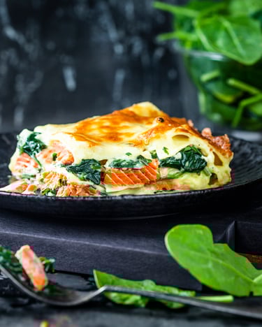 Vor dunklem Hintergrund sticht die Lachs-Spinat-Lasagne auf dem schwarzen Teller farblich hervor. Knusprig gebackener Käse und die Schichten von Nudeln, Füllung und Soße sehen einladend lecker aus.