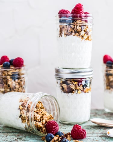 Granola mit griechischem Joghurt und Früchten in Schraubgläsern, neben Holzlöffelchen.