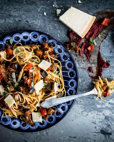 Spaghetti mit Auberginen-Paprika-Pesto auf eine blauen Teller, daneben ein Stück Parmesan.