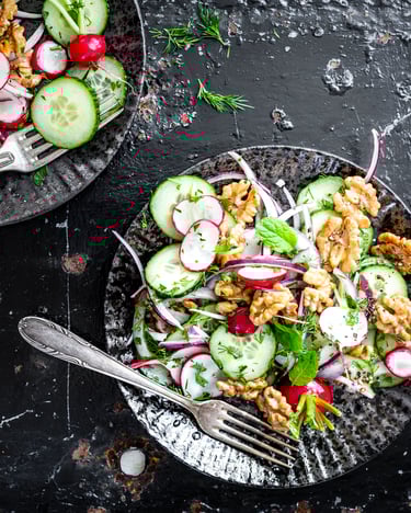 Salat aus Gurken, Walnüssen, Radieschen und roten Zwiebeln auf schwarzen Tellern.