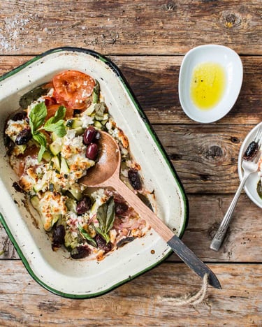 Feta mit Oliven, Tomaten und Zucchini in einer ofenfesten Form gratiniert mit Olivenöl und Besteck auf Holzuntergrund.