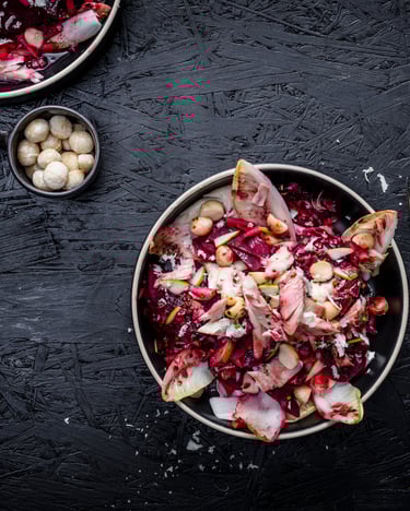 Rote-Bete-Salat mit Pflaumenvinaigrette und gezupfter Räucherforelle, angerichtet auf einem Teller, von oben fotografiert. Daneben eine Gabel, in einer kleinen Schüssel liegen Macadamianusskerne. Alles auf dunklem Untergrund.