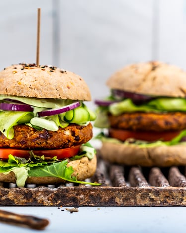 Man sieht zwei Burger, die mit einem Bohnenpatty, Gurken, Zwiebeln, Salat und Tomate belegt sind