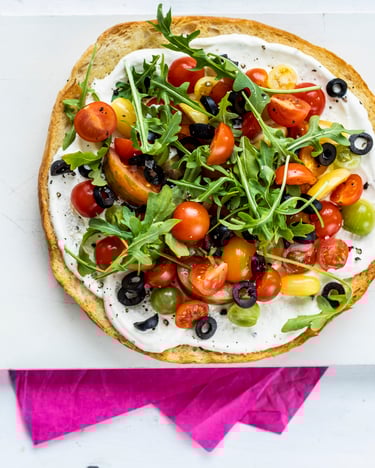 Längs halbiertes Fladenbrot mit Schmand, Tomaten, Oliven und Rucola belegt auf einem weißen Brett mit einer pinken Serviette.