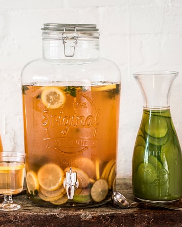 In einem durchsichtigen, großen Bowle-Glas schwimmt Eistee, orange, mit Limetten und Zitronenscheiben – daneben weitere Erfrischungsgetränke. Fotografiert von vorne vor einer weißen Wand.