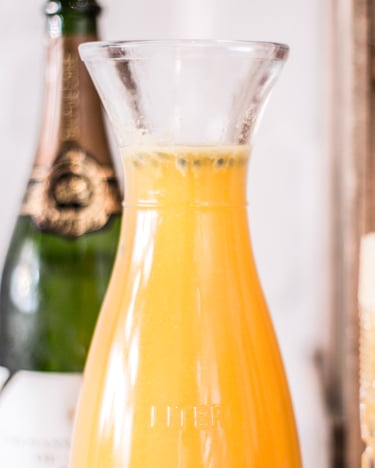 Eine Karaffe mit gelbem Fruchtmix-Mango-Maracuja-Drink neben zwei Gläsern mit Strohhalm.