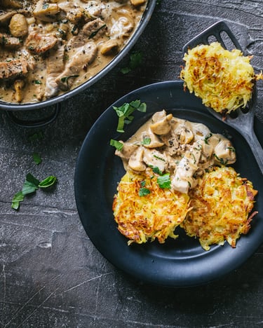 Topshot von Zürcher Geschnetzeltem mit Champignons und Kartoffelrösti auf schwarzem Teller und dunklem Untergrund
