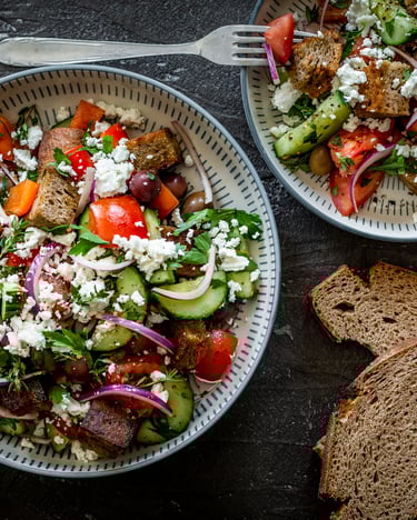 Zwei Teller mit buntem Brotsalat und angelegtem Bauernbrotscheiben auf grauem Untergrund