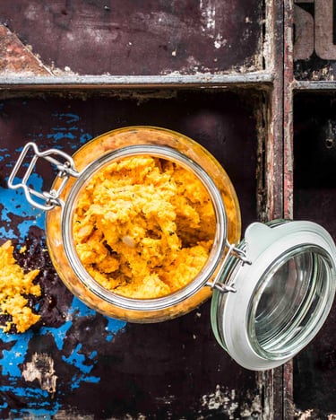 Möhrenhummus in einem Weckglas mit offenem Deckel, daneben ein Löffel mit etwas Hummus.