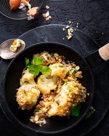 Blumenkohl mit Polonaise und Schinken-Béchamel, fotografiert in einer kleinen, dunklen Pfanne auf dunklem Untergrund, garniert mit Petersilie. Etwas Eierschale liegt im Bild.