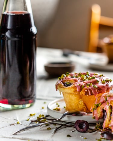 Hefeschnecken mit Sauerkirschen aus der Muffinform, mit pinker Sauerkirschglasur und knackigen, grünen Pistazienstücken, auf weißem Teller mit einer Sauerkirschnektarflasche im Bild.