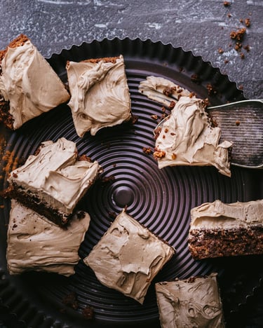 Stücke von Carob-Brownies auf einer dunklen Servierplatte, ein Stück liegt auf einer Kuchenschaufel. Fotografiert von oben auf dunklem Untergrund, etwas Carob-Pulver ist noch verstreut.