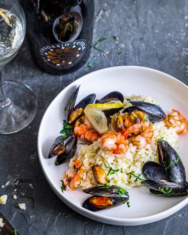 Risotto di mare mit Garnelen und Muscheln auf weißem Teller und dunklem Untergrund, dazu ein Glas Prosecco im Hintergrund