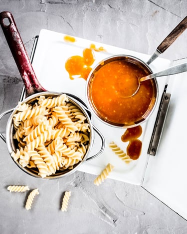 Fusilli und Tomatensauce jeweils in kleinen Töpfen auf einem Miniaturherd und grauem Untergrund stehend.