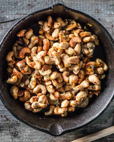Geröstete Cashewkerne mit Gewürzen in einer gusseisernen Pfanne auf Holzuntergrund.