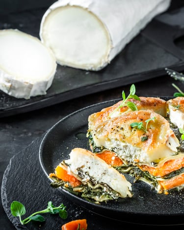 Kürbis-Spinatlasagne mit Ziegenkäse auf einem dunklen Teller, seitlich fotografiert, im Hintergrund große Ziegenkäserolle.