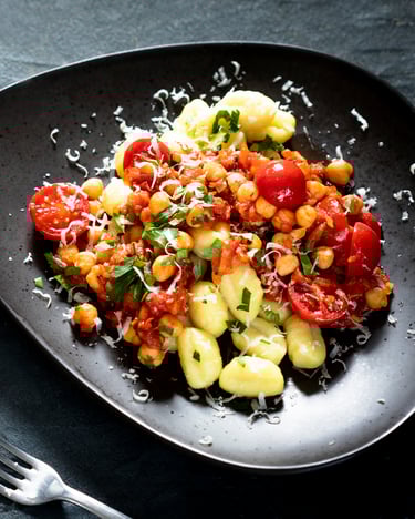 Gnocchi mit Tomatensauce und Kichererbsen auf dunklem Teller, daneben ein Schälchen Parmesan