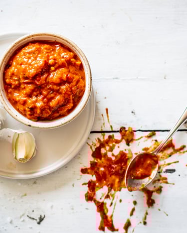Roter Tomaten-Paprika-Dip in Schale und auf weißem Holztisch gekleckert mit Knoblauchzehe und Löffel.