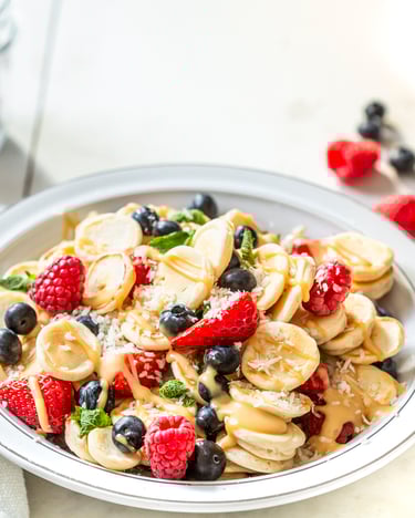 Pancake Cereals – Mini-Pfannkuchen in einer Frühstücksschüssel mit roten Beeren, Mandelmus und Minze, fotografiert seitlich auf hellem Untergrund.