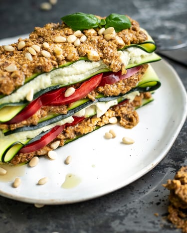 Ein Stück Lasagne mit Schichten aus Zucchinistreifen, Paprikaringen, Cashew-Käse und Tomaten-Pesto getoppt mit Pinienkernen auf weißem Teller