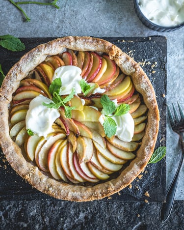 Der Apfelkuchen, gedeckt mit Apfelscheiben, drei Klecksen Joghurt und Minzblätterb, steht auf einem schwarzen Tafelbrett. Ein paar Minzblätter und Gabeln liegen daneben auf einem grauen Tisch.