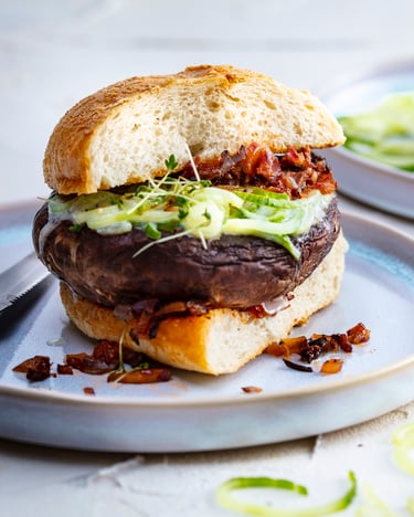 Veganer Portobello-Burger mit Gurken-Spaghetti-Füllung und Ciabattabrot, fotografiert von vorne auf hellen Tellern, der Burger ist angeschnitten, neben dem Teller liegen ein paar Gurken-Spaghetti.