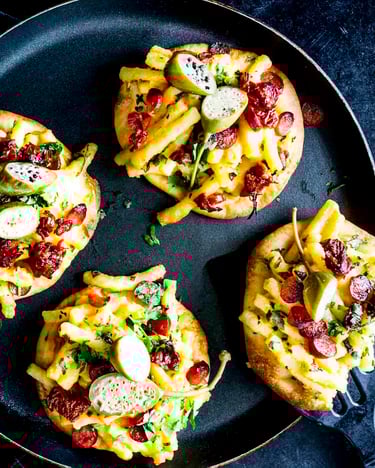 Vier Mini-Pizzas mit Belag aus Mac’n’Cheese und getrockneten Tomaten auf schwarzem Teller und schwarzem Grund
