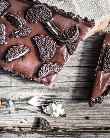 Zerteilte Oreo-Kekse auf einem No Bake Käsekuchen mit Schokoladen-Ganache auf Holzuntergrund.