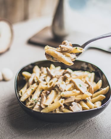 Pilzragout mit Steinpilzen auf dunklem Pastateller fotografiert auf hölzernem Untergrund. Pilze liegen an den Seiten, eine Hand hält einen Löffel mit etwas Pasta kurz über dem Teller.