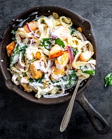 Pfanne mit Fettuccine, Sahnesoße, Lachs und frischem Spinat auf schwarzem Tisch.