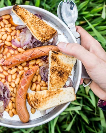 Baked Beans mit Würstchen, roten Zwiebeln und Toast in einem Campinggeschirrteller mit Löffel.