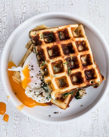 Käse-Spinat-Waffeln mit pochiertem Ei, zwei Waffeln mit Ei dazwischen auf einem weißen Teller, fotografiert von oben auf hellem Untergrund.
