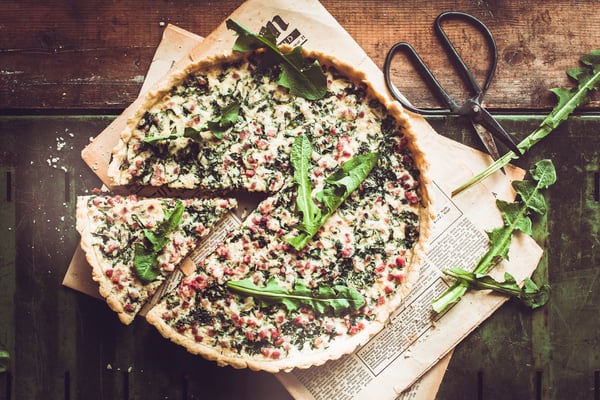 Angeschnittene Tarte mit Speck und Löwenzahn auf Holzuntergrund mit angelegter Kräuterschere