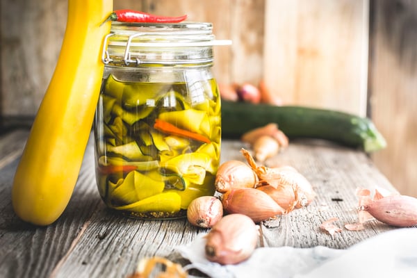 Eingelegte Zucchinistreifen mit Chilis in Einmachglas auf Holz neben gelber und grüner Zucchini und Zwiebeln